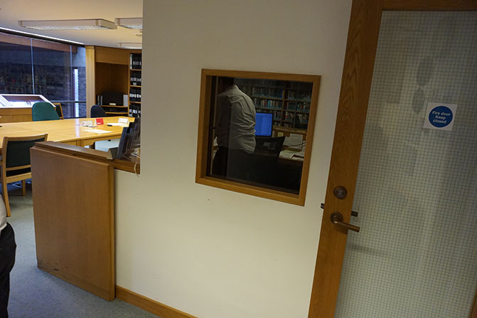 View from the staff desk in the Archives Centre reading room. There is a window into a small room (the microfilm reading room)