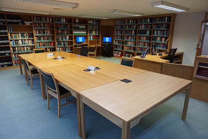 The Archives Centre reading room post-refurbishment. The same large table stands in the middle of the room. Beyond it, the staff desk has been extended and there are more bookshelves, and 2 computer work stations.