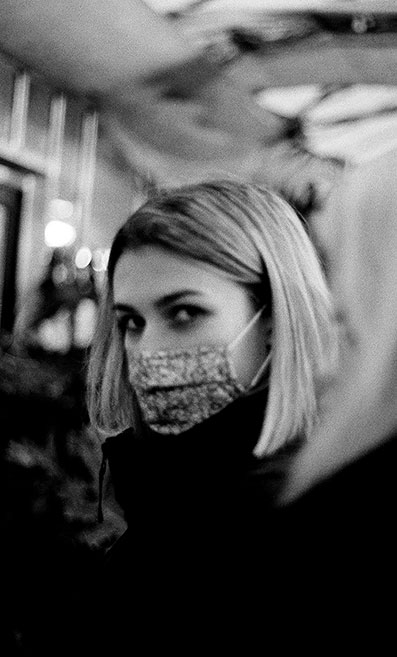 A black and white photo of a young woman wearing a face covering, looking directly into the camera.