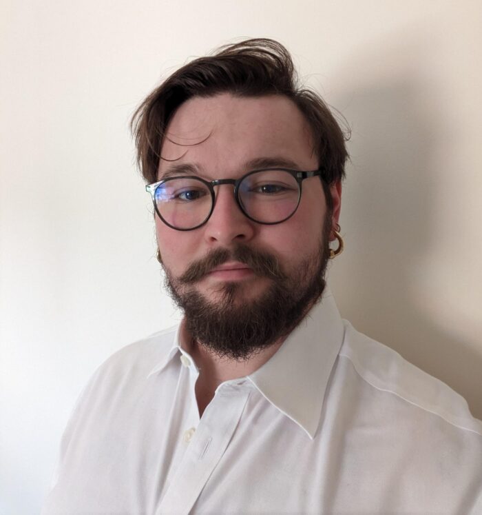 Colour head and shoulders photograph of Anglia Ruskin student Archie Jennings, wearing a white shirt and standing in front of a cream wall