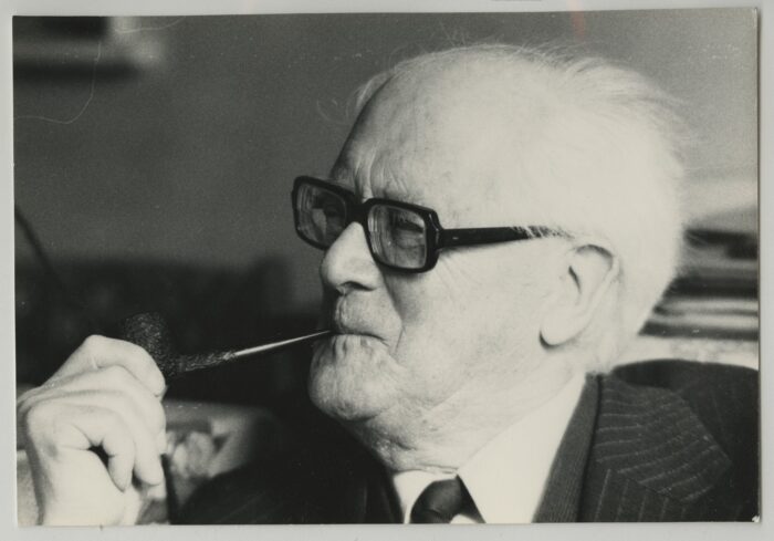 Black and white head and shoulders photograph of Fenner Brockway sitting on a sofa and smoking a pipe