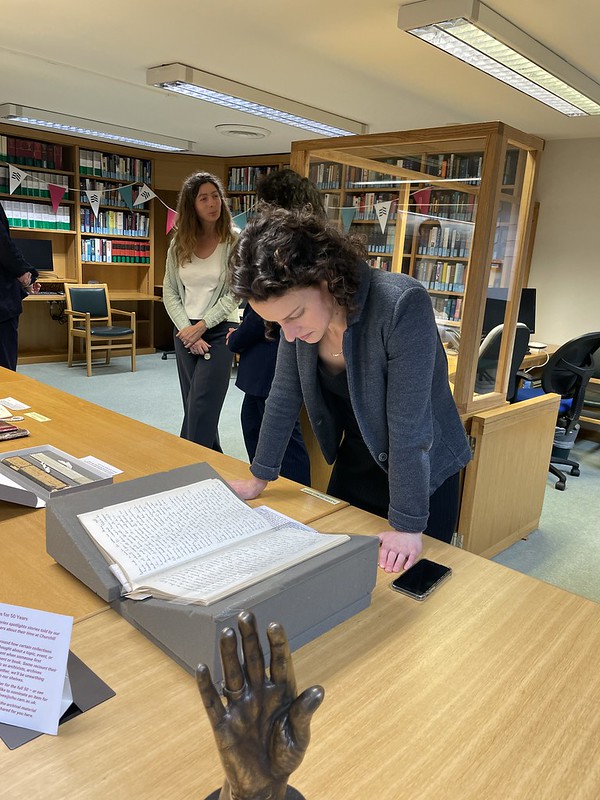 A person with curly hair is deeply engrossed in a large open book on a table in the library, perhaps exploring grants and by-fellowships. Other patrons peruse shelves in the background, while a bronze sculpture of a hand subtly adorns the table.