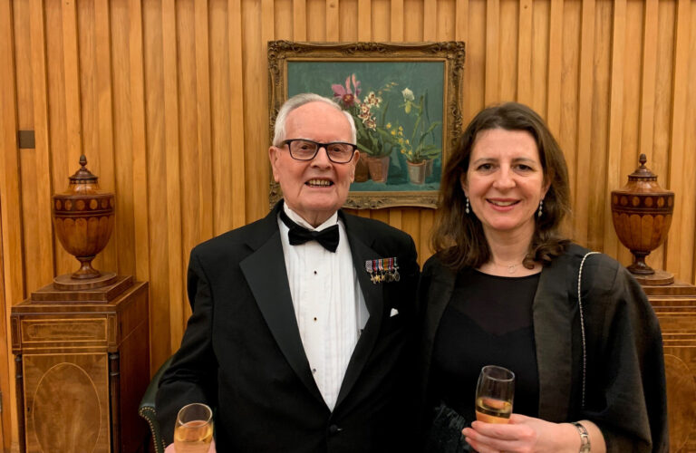 An elderly man in a tuxedo and bow tie stands beside a woman with long brown hair in an elegant black dress. Both are holding champagne glasses, smiling, and celebrating the announcement of prestigious grants in a warmly lit room with wooden walls and framed artwork.