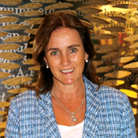 A person with long brown hair smiles at the camera, wearing a blue checkered blazer and a white top. They stand before a decorative backdrop featuring abstract text and patterns, reminiscent of artistic grants and fellowships promoting creative expression.