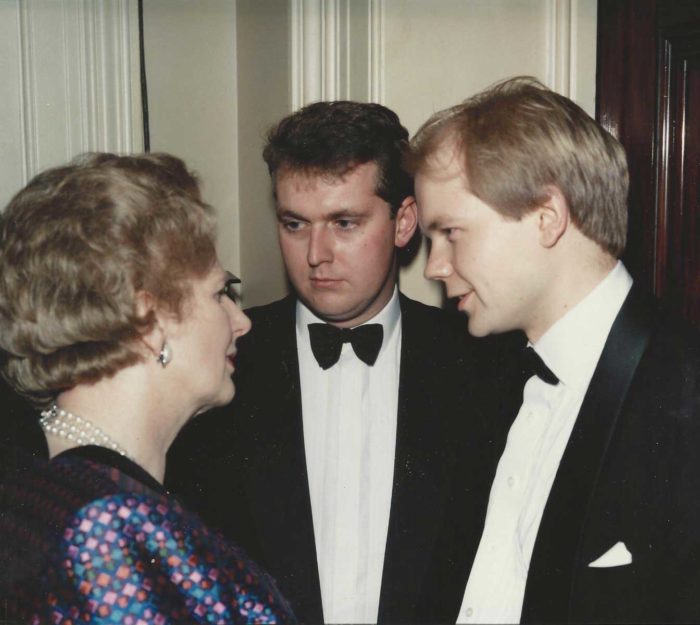 Three people in formal attire are engaged in a conversation about grants. An older woman with short hair and glasses faces two younger men wearing tuxedos. They stand in a warmly lit room with cream-colored walls, where discussions about prestigious by-fellowships often take place.