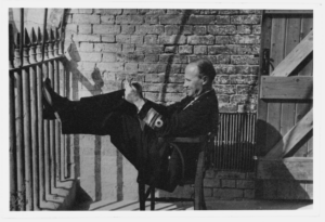 A black and white photograph: a man in uniform sits on a chair with his feet propped up at shoulder height against an iron railing