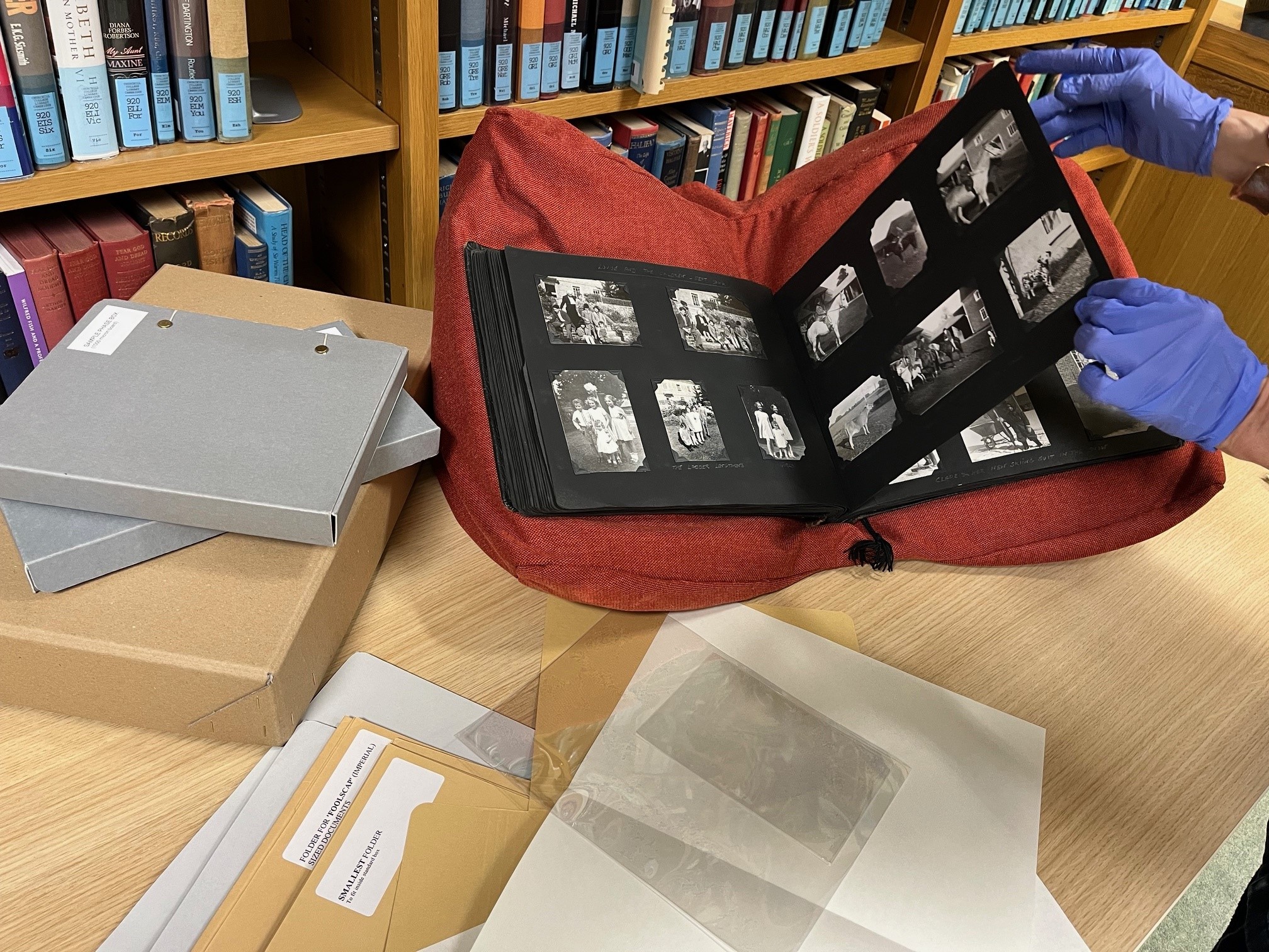 Photograph album on a bookrest with pages being turned by gloved hands. Surrounded by a selection of packaging materials